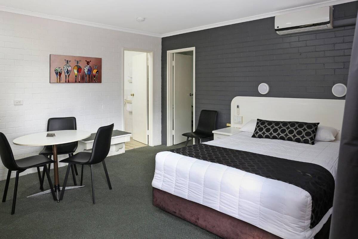 A hotel room with a bed, a desk with chairs, a painting on the wall, and an air conditioning unit.
