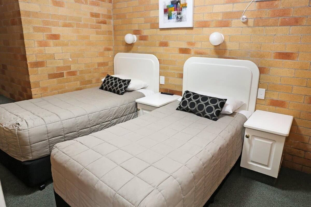 A room with two beds, each with a white headboard and a grey quilted bedspread. There are two bedside tables with lamps, and a small cabinet with a lock. The walls are brick, and there is a framed picture on the wall.