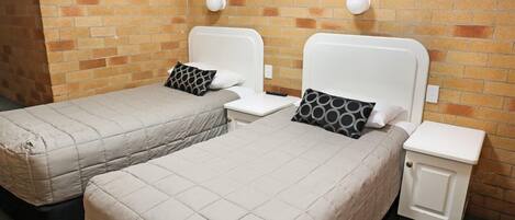 Una habitación con dos camas, cada una con cabecero blanco y colcha acolchada gris. Hay dos mesitas de noche con lámparas, y un mueble pequeño con cerradura. Las paredes son de ladrillo, y hay un cuadro en la pared.