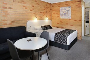 Una habitación de hotel con pared de ladrillo, cama con colcha estampada, cabecero blanco, sofá negro y una pequeña mesa redonda con un letrero.