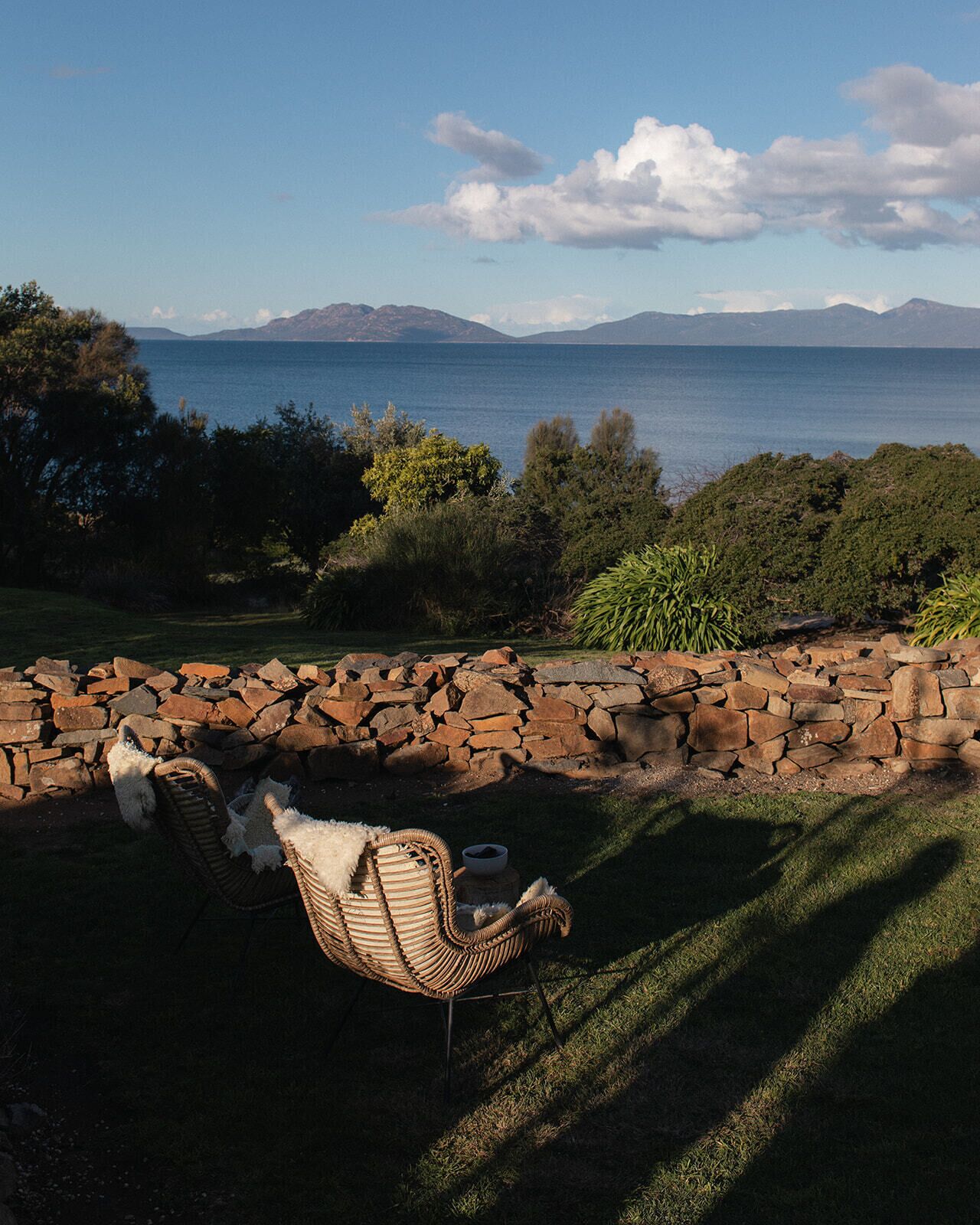 A wicker chair with a cushion and a bowl on a stone wall overlooking a body of water with mountains in the distance.