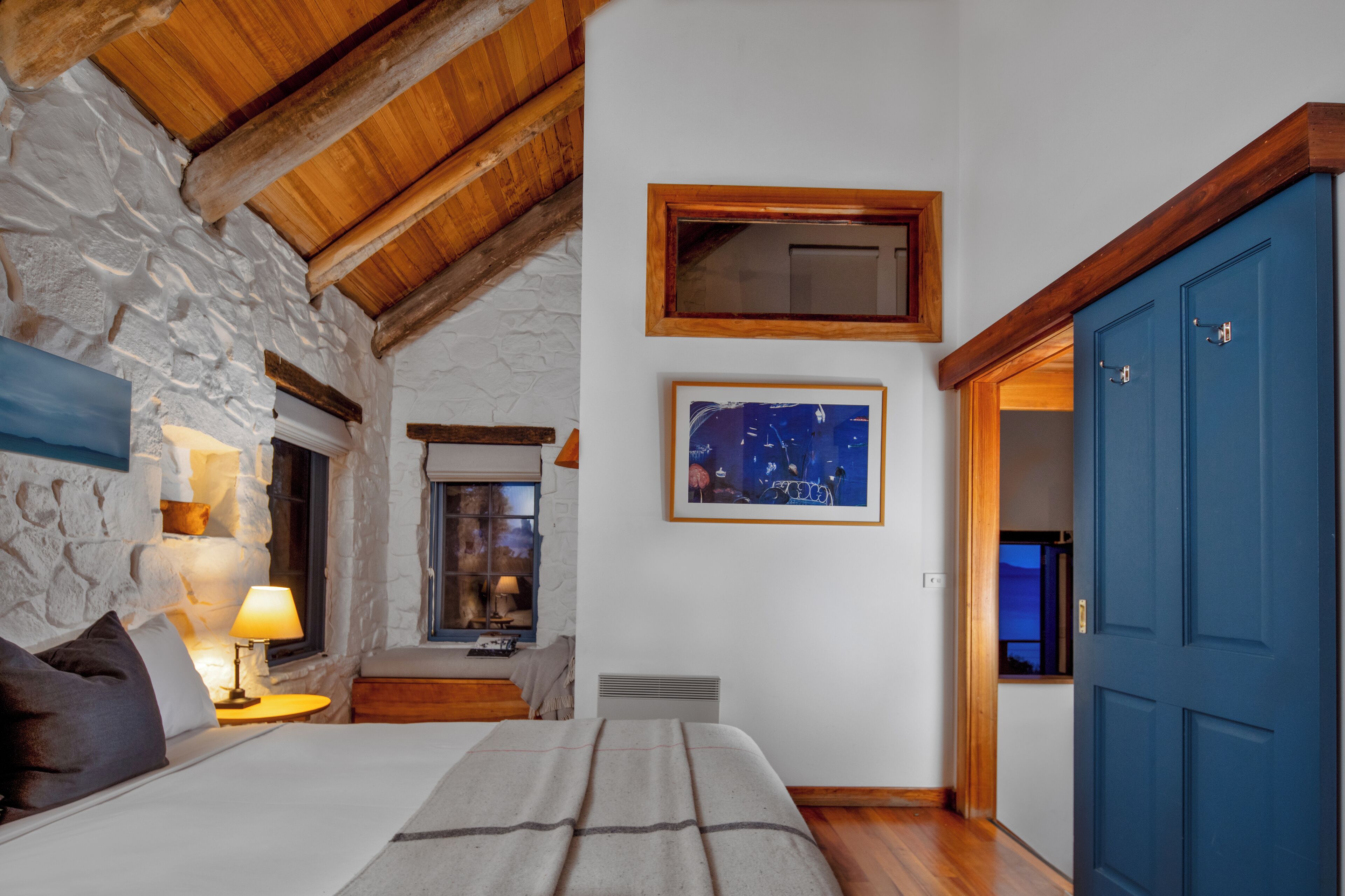 A cozy bedroom with a stone wall, wooden beams, a bed with a blanket, a nightstand with a lamp, and a framed picture on the wall.