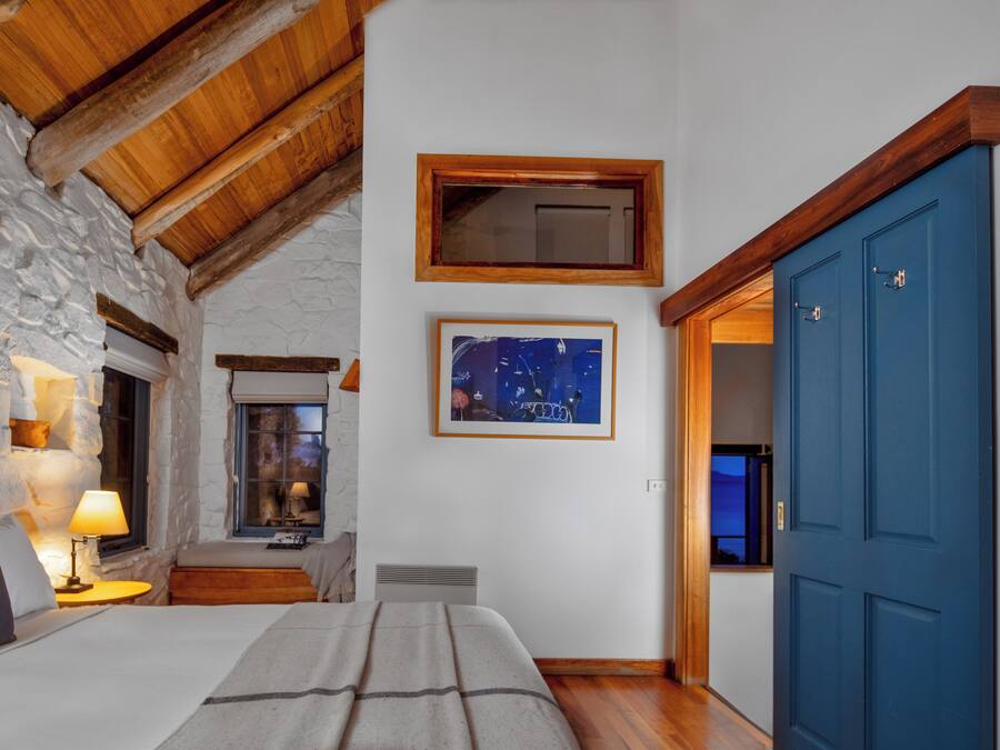 A cozy bedroom with a stone wall, wooden beams, a bed with a blanket, a nightstand with a lamp, and a framed picture on the wall.