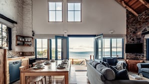 Een ruime woonkamer met een grote houten eettafel, een comfortabele bank en uitzicht op zee door openslaande deuren.