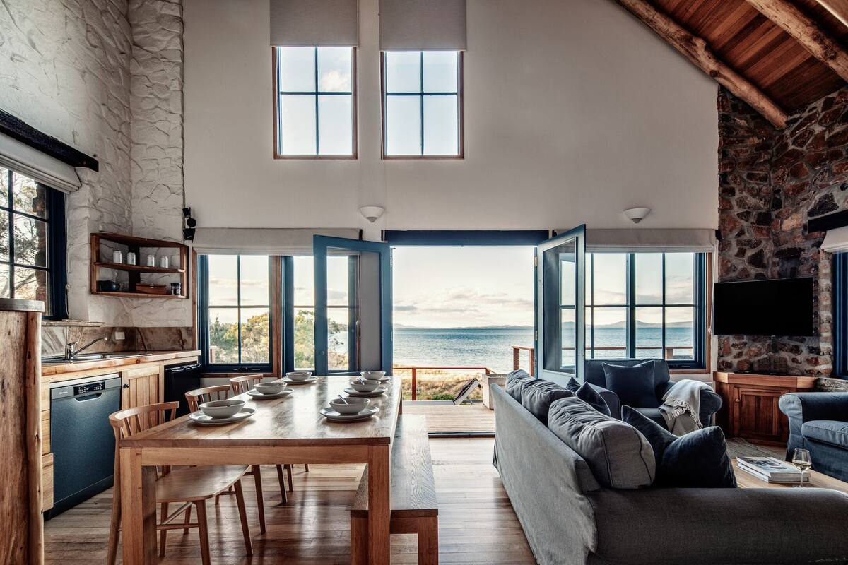 A spacious living area with a large wooden dining table, a comfortable sofa, and a view of the sea through open doors.
