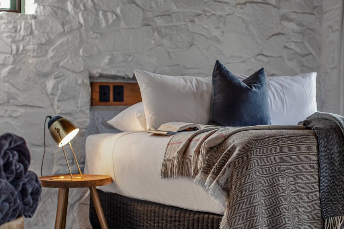 A bedroom with a stone wall, a bed with white and dark pillows, a wooden headboard, a bedside lamp, and a woven basket.