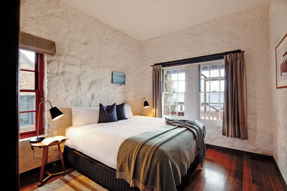 A bedroom with a large bed, a bedside table with a lamp, a window with curtains, and a stone wall.