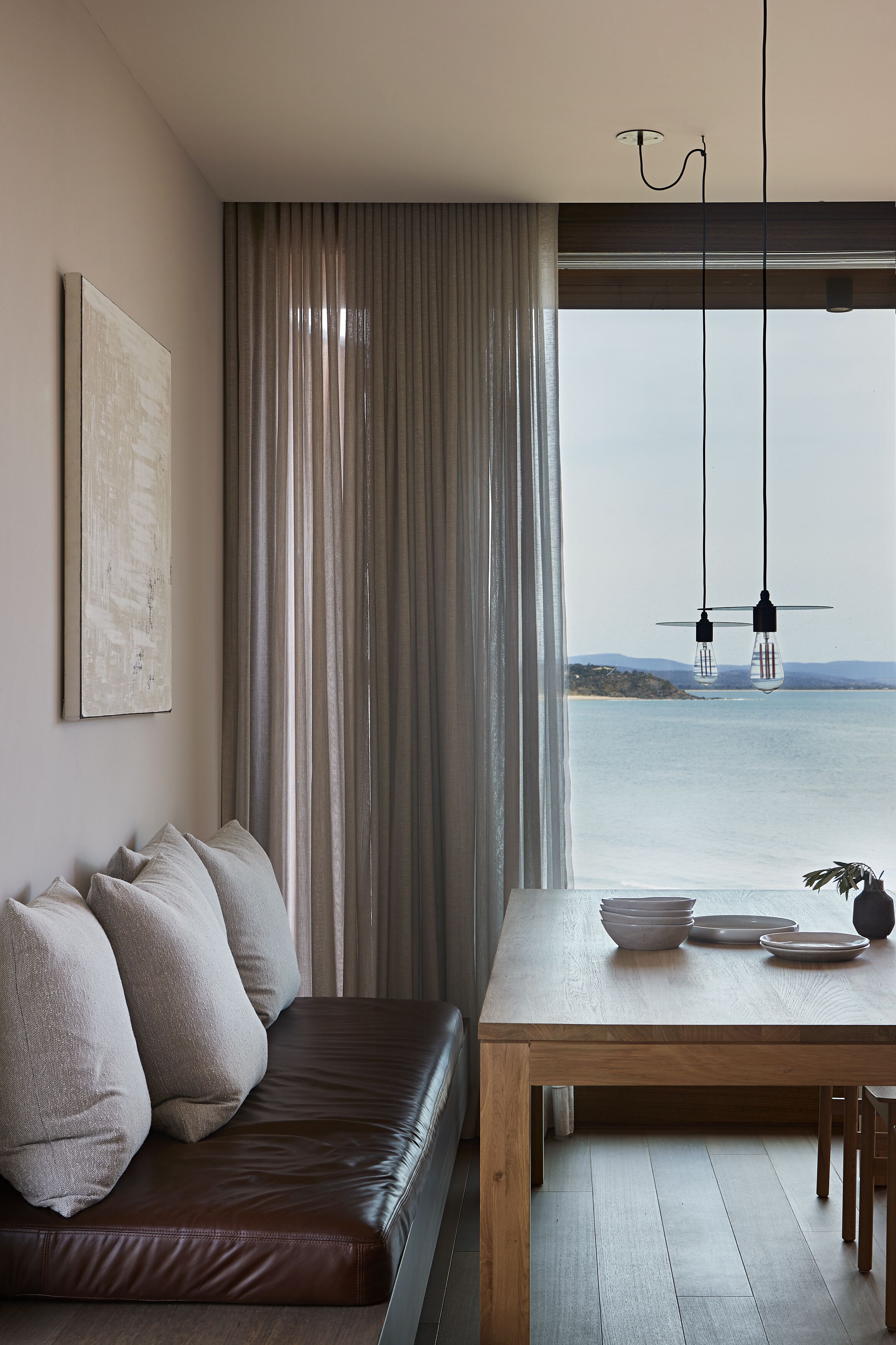 A modern living room with a leather sofa, a wooden dining table, and a view of the sea.