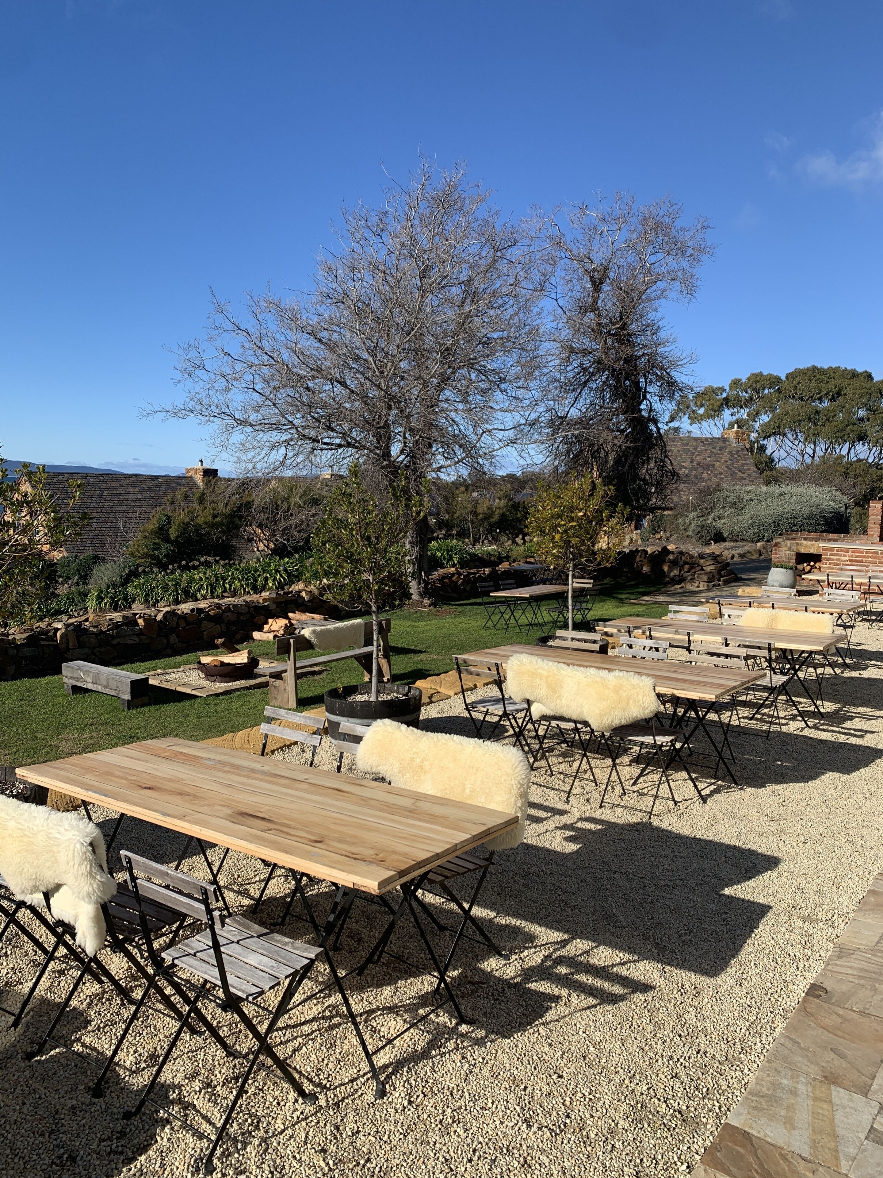 An outdoor seating area with wooden tables and chairs, surrounded by gravel and greenery.