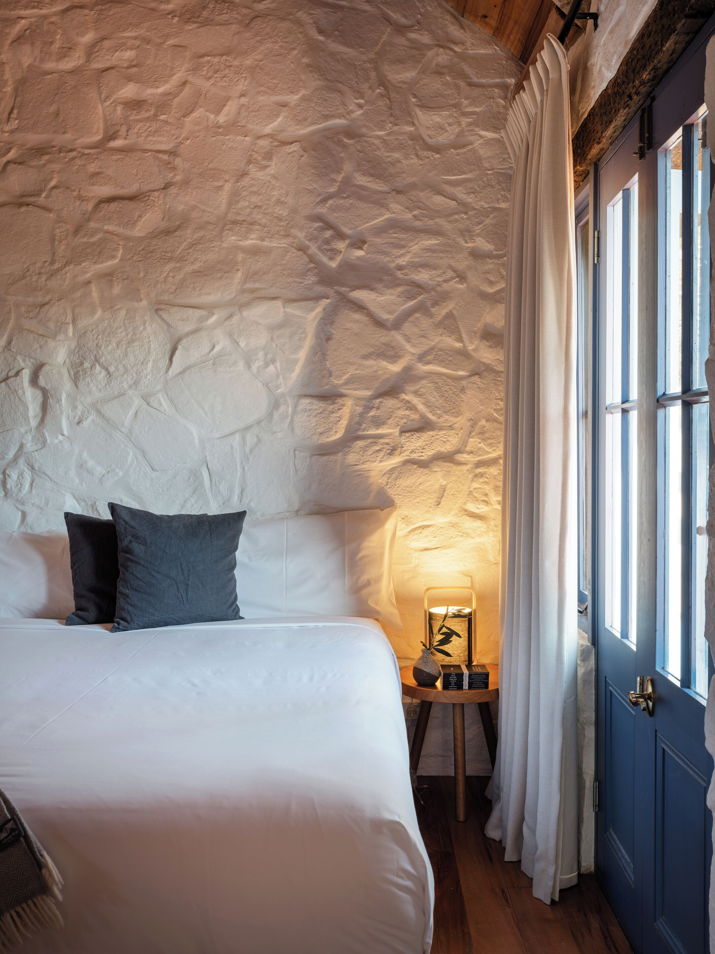 Un dormitorio con pared de piedra, una cama con ropa blanca, dos almohadas, una mesita de noche con lámpara y una puerta azul.