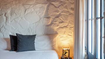 Un dormitorio con pared de piedra, una cama con ropa blanca, dos almohadas, una mesita de noche con lámpara y una puerta azul.