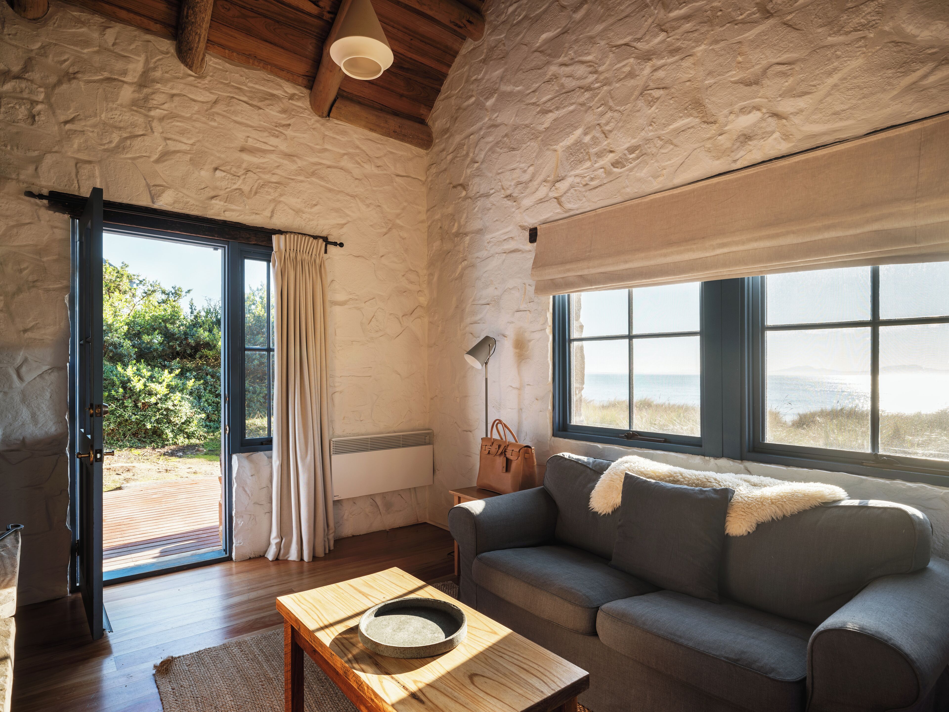 A cozy room with a stone wall, a large window with a view, a grey sofa, a wooden table, and a door leading outside.