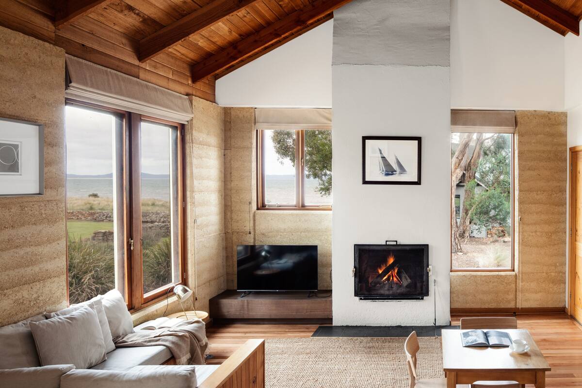 A spacious living room with a large TV, fireplace, and multiple windows offering views of the outdoors.