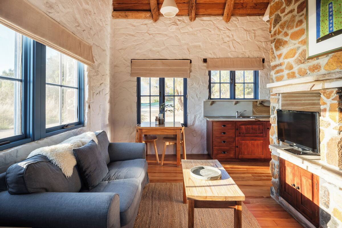 A cozy living room with a sofa, a dining table, and a kitchen area with a stone wall and wooden beams.