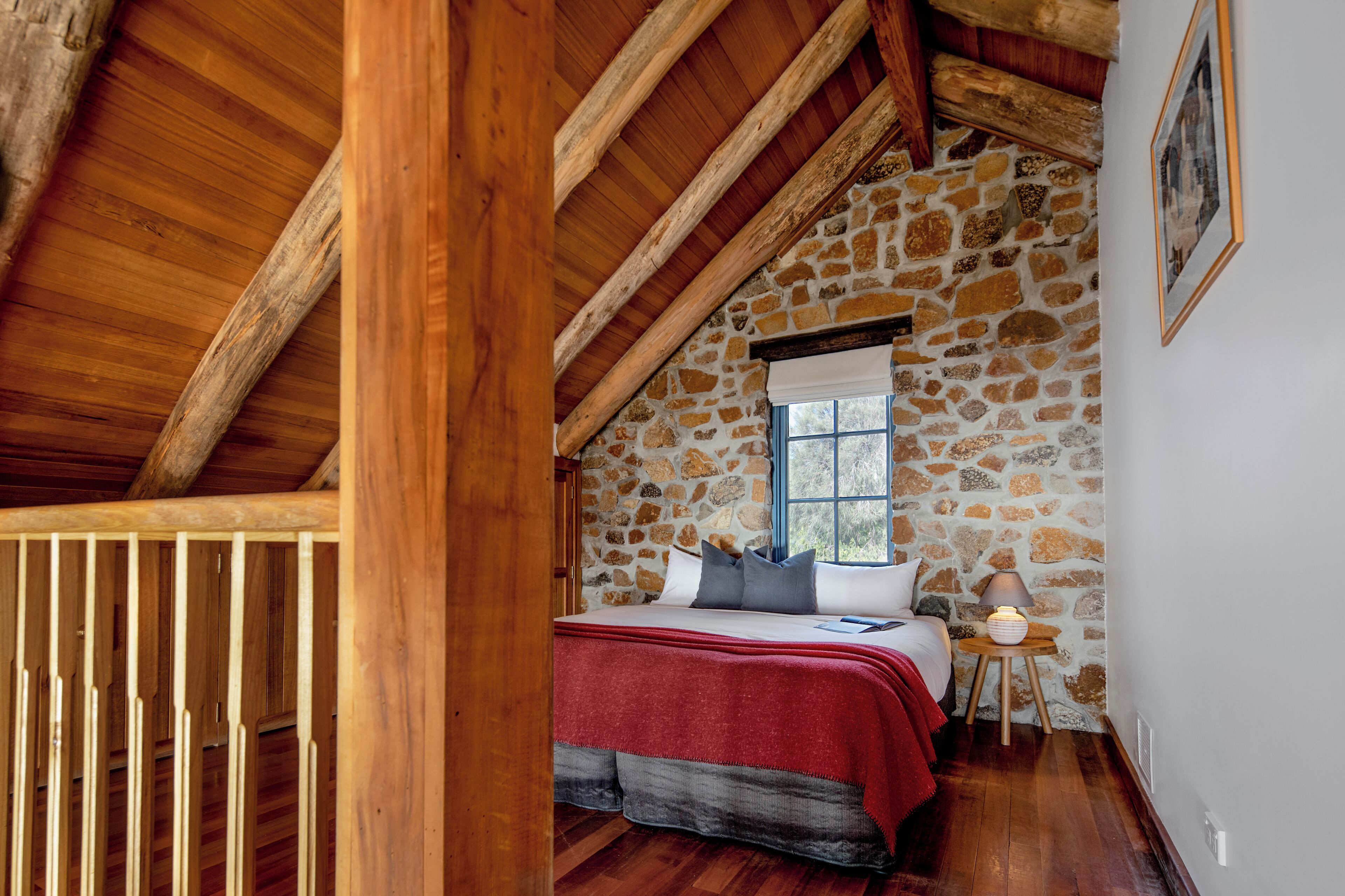 Une chambre avec un mur en pierre, des poutres en bois, un lit recouvert d’une couverture rouge, une fenêtre et une table de chevet avec une lampe.