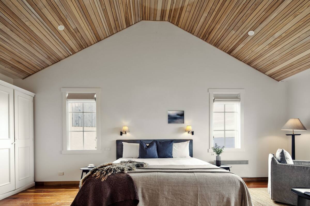 A bedroom with a sloped wooden ceiling, a bed with a grey and white headboard, two bedside lamps, a window, and a grey sofa.
