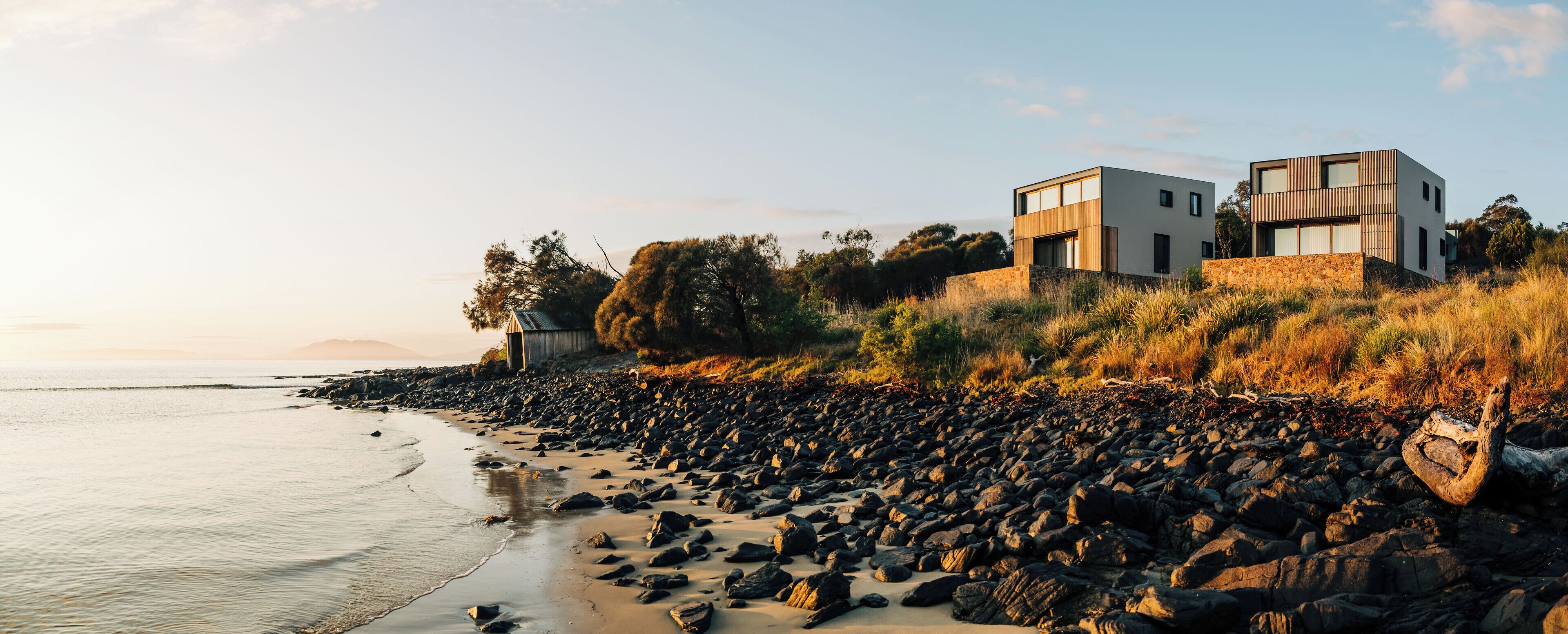 Une propriété moderne située en bord de mer, offrant une vue imprenable sur la plage, et comprenant un bâtiment à deux étages avec des fondations en pierre et des éléments décoratifs en bois.
