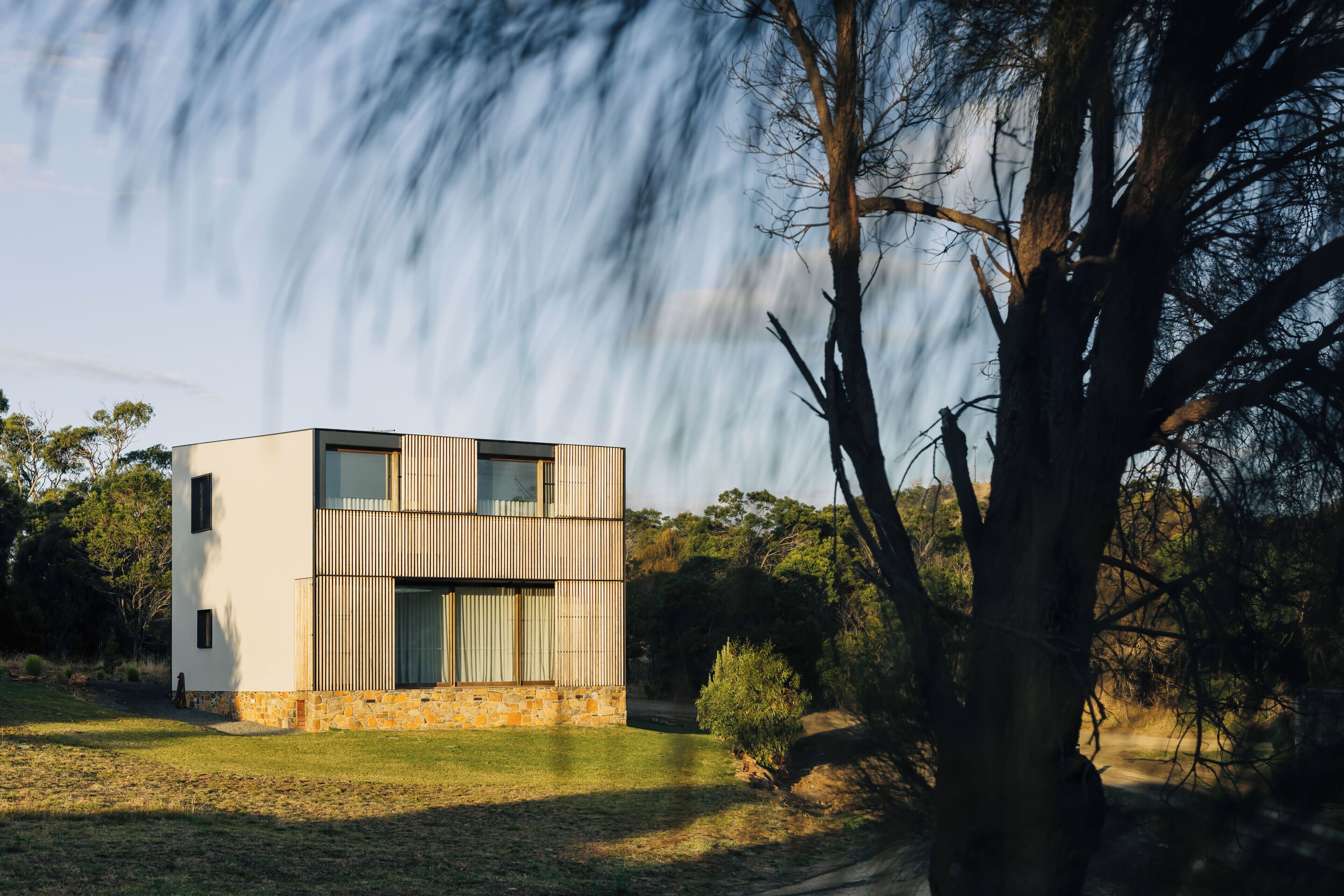 A modern two-story house with a stone foundation, large windows, and a surrounding natural landscape.