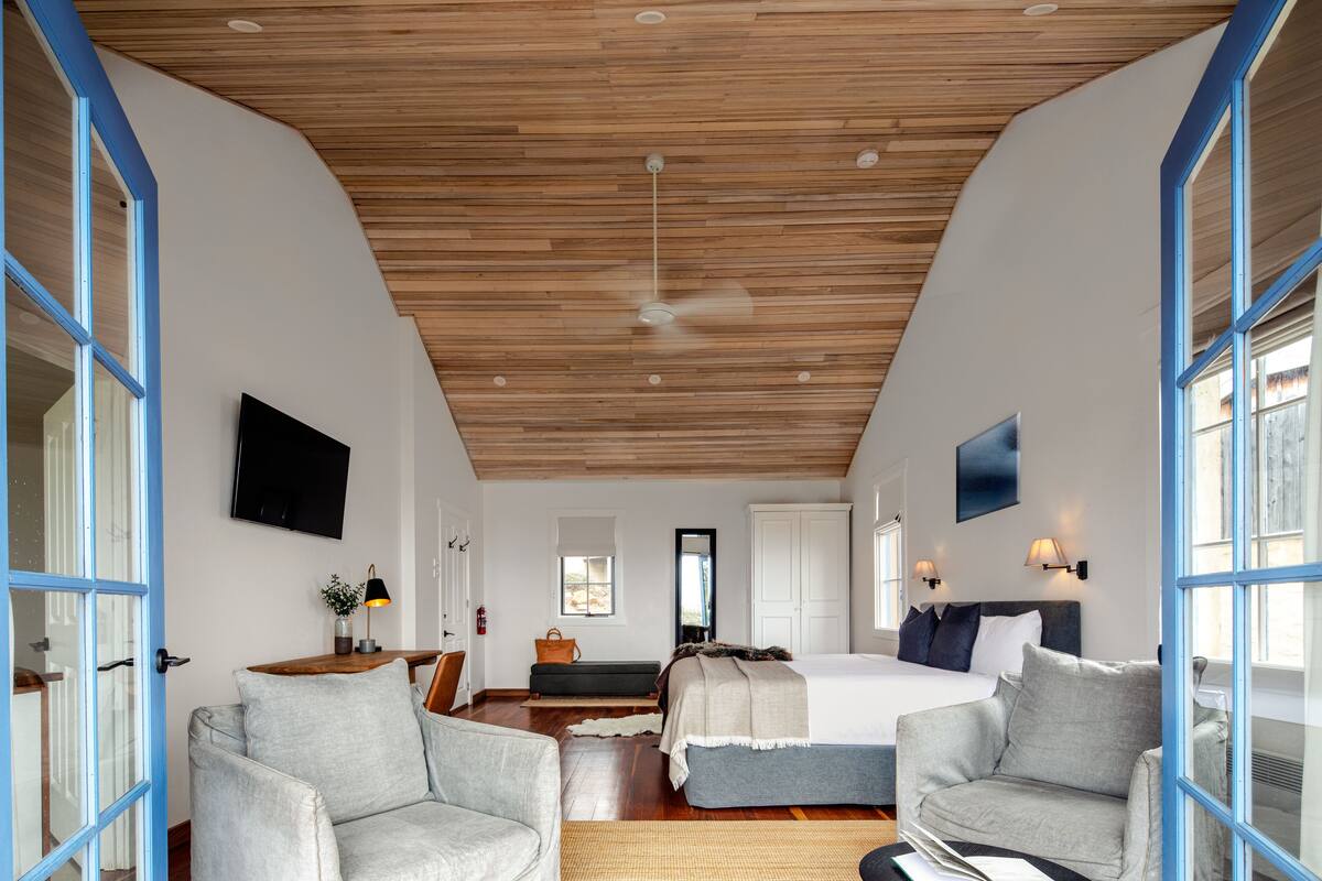 A spacious bedroom with a wooden ceiling, a large bed, two armchairs, and a desk with a lamp.
