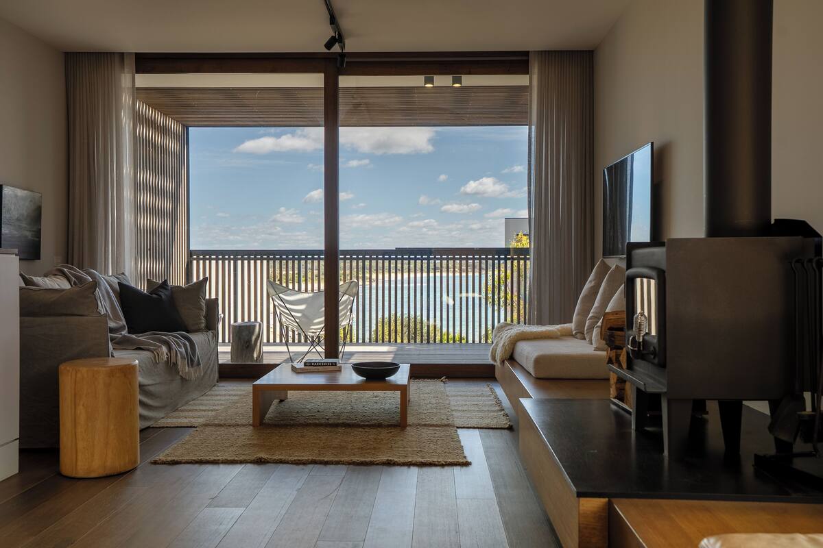 A modern living room with a sofa, coffee table, and TV. Large windows offer a view of the outdoors.