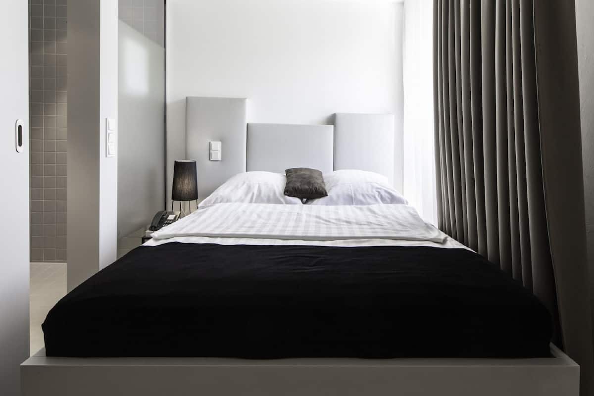 A modern hotel room with a large bed, a bedside table with a lamp, and a view of the outdoors.