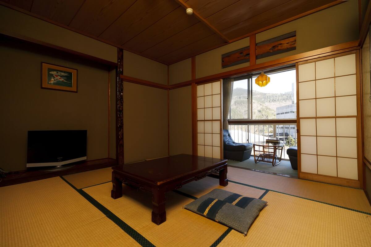Una habitación japonesa tradicional con suelo de tatami, una mesa de madera baja, un televisor y una puerta corredera que da a un balcón.