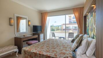 Una habitación de hotel con cama, escritorio, silla, banco y un amplio ventanal con cortinas.