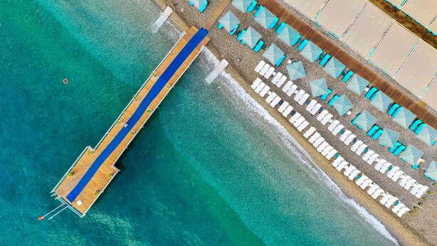 An aerial view of a beach with a wooden pier extending into the water, lined with rows of white lounge chairs and blue umbrellas.