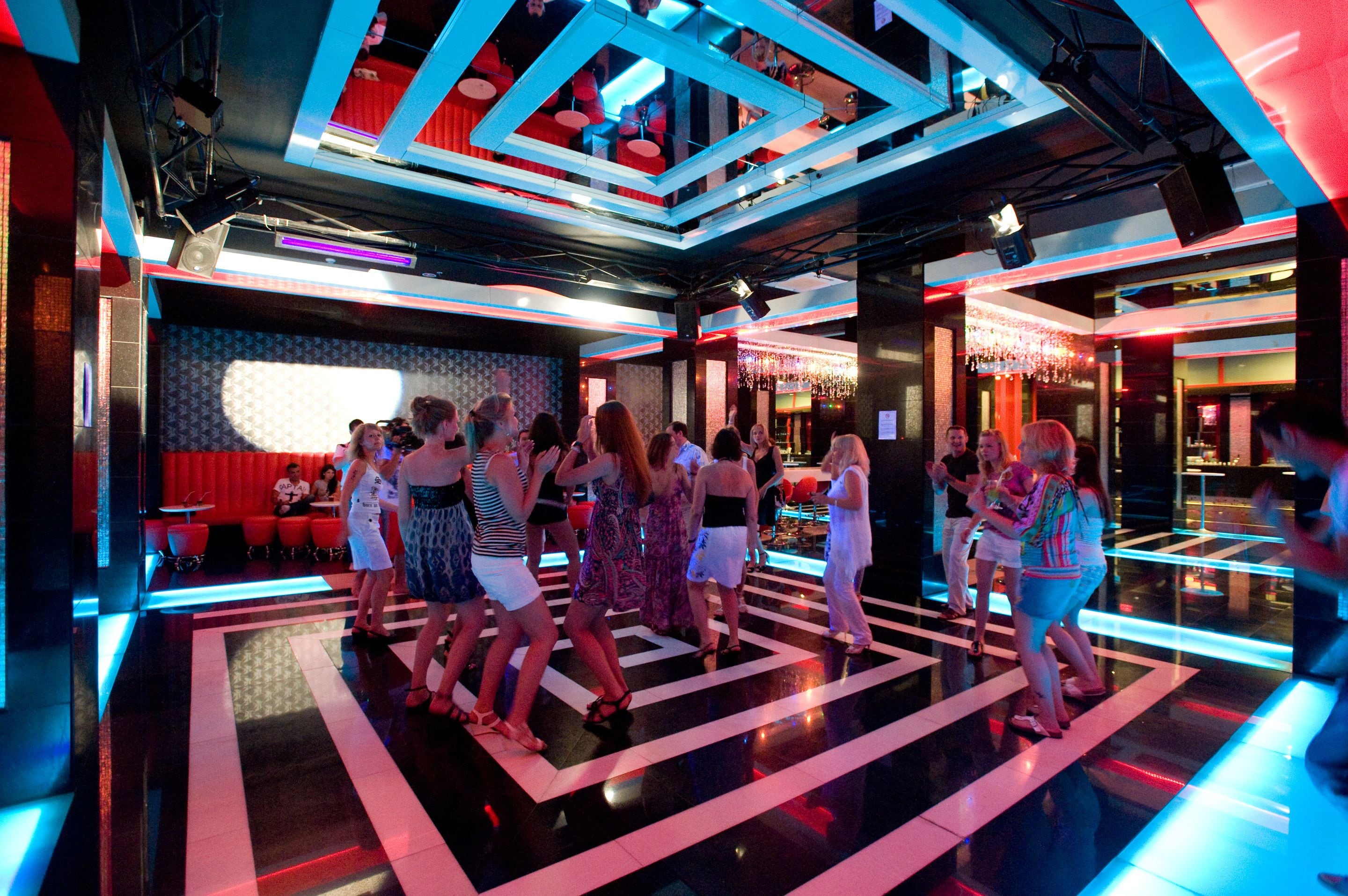 A dance floor with people dancing in a brightly lit room with geometric ceiling designs and a bar area in the background.