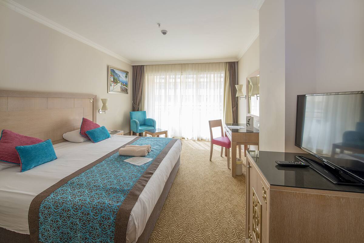 Une chambre d’hôtel avec un grand lit, un bureau, une chaise, une télévision et une fenêtre avec des rideaux.