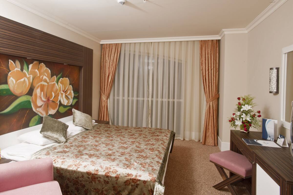 Une chambre d’hôtel avec un lit, un bureau, une chaise, un tableau et une fenêtre avec des rideaux.