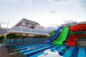 Una piscina con múltiples toboganes, un área de descanso techada y un edificio con balcones al fondo.