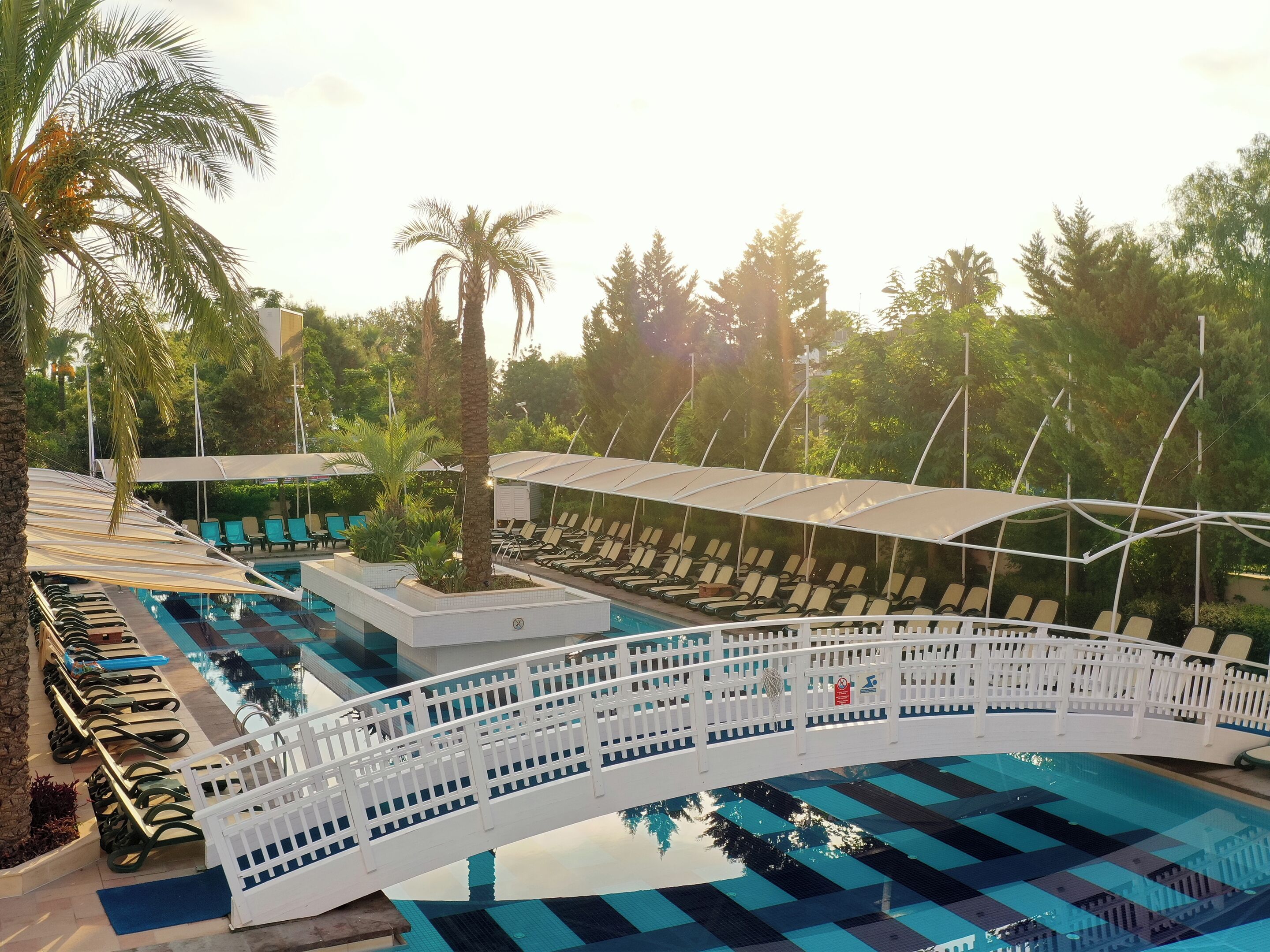 Un área de piscina con sillas de descanso, un puente blanco y palmeras.