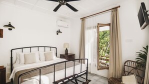 A bedroom with a bed, a ceiling fan, an air conditioner, a nightstand, a lamp, a wicker chair, and a window with curtains. - Brown Feather Batu Belig (Keroboken Kelod)