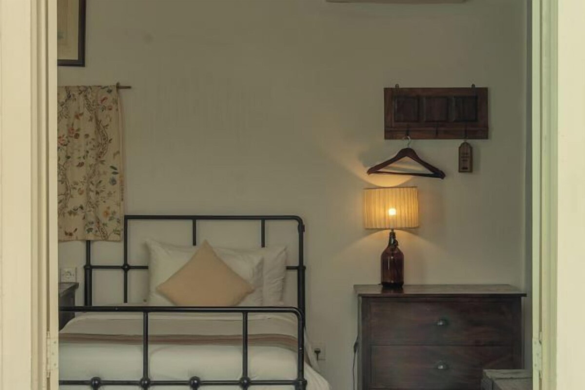 Una cama individual con estructura metálica, una mesita de noche con lámpara y un aparador de madera.