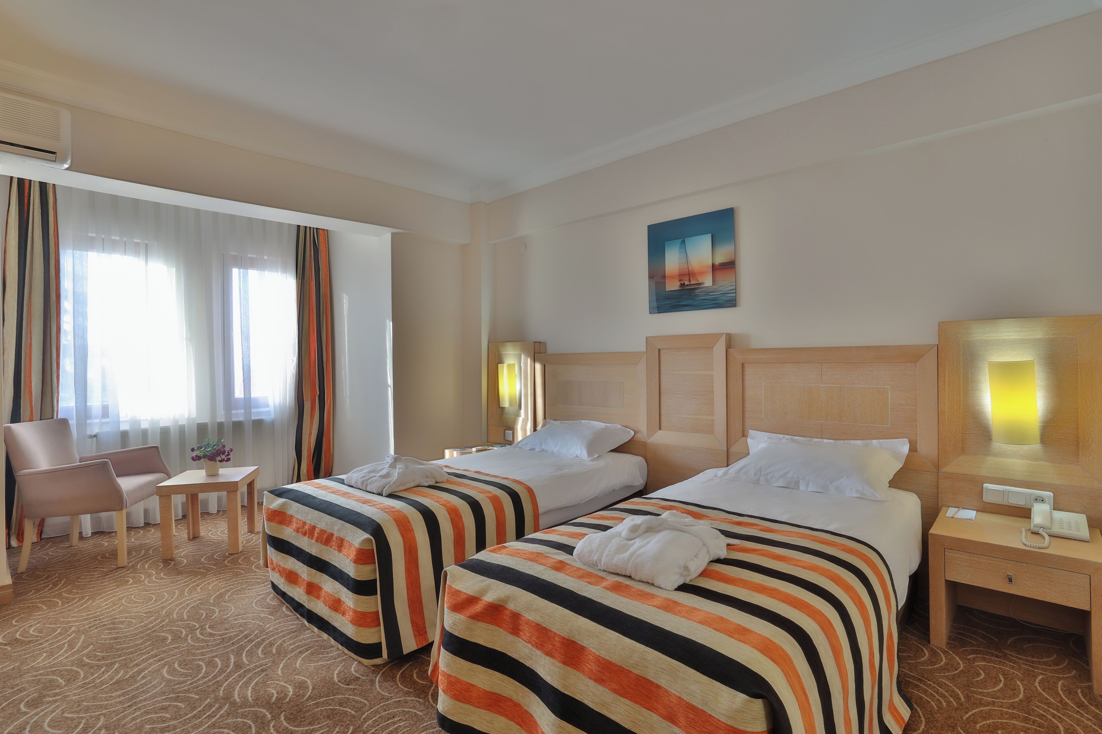 A hotel room with two beds, a chair, a small table, and a nightstand with a phone.