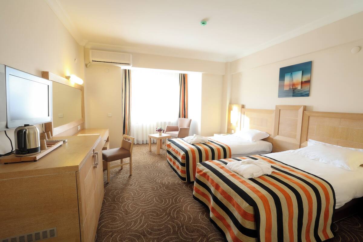 A hotel room with two beds, a desk with a kettle, a chair, a small table with a vase, and a window with curtains.