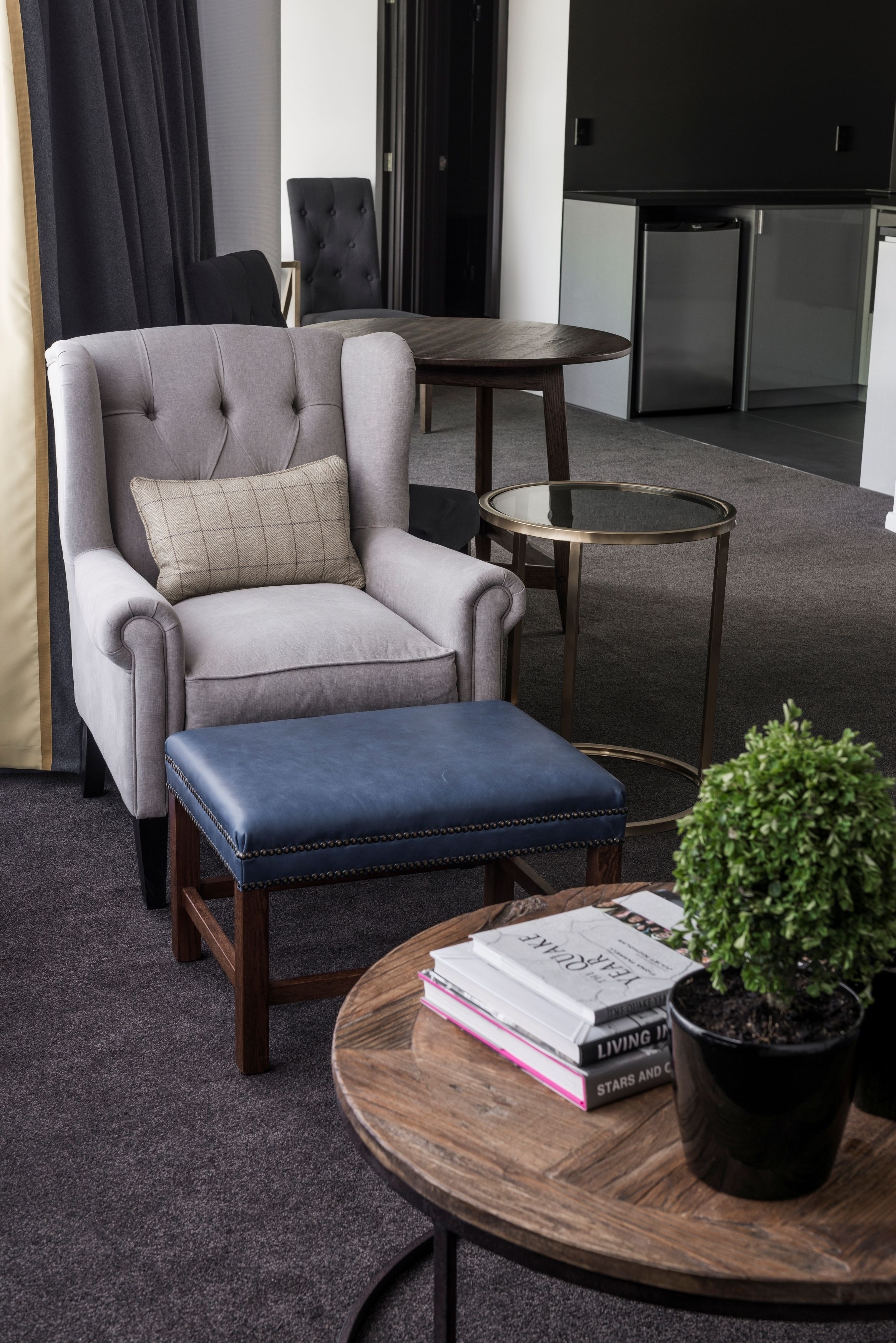 A modern living room with a grey armchair, a blue ottoman, a round wooden coffee table with books and a potted plant.