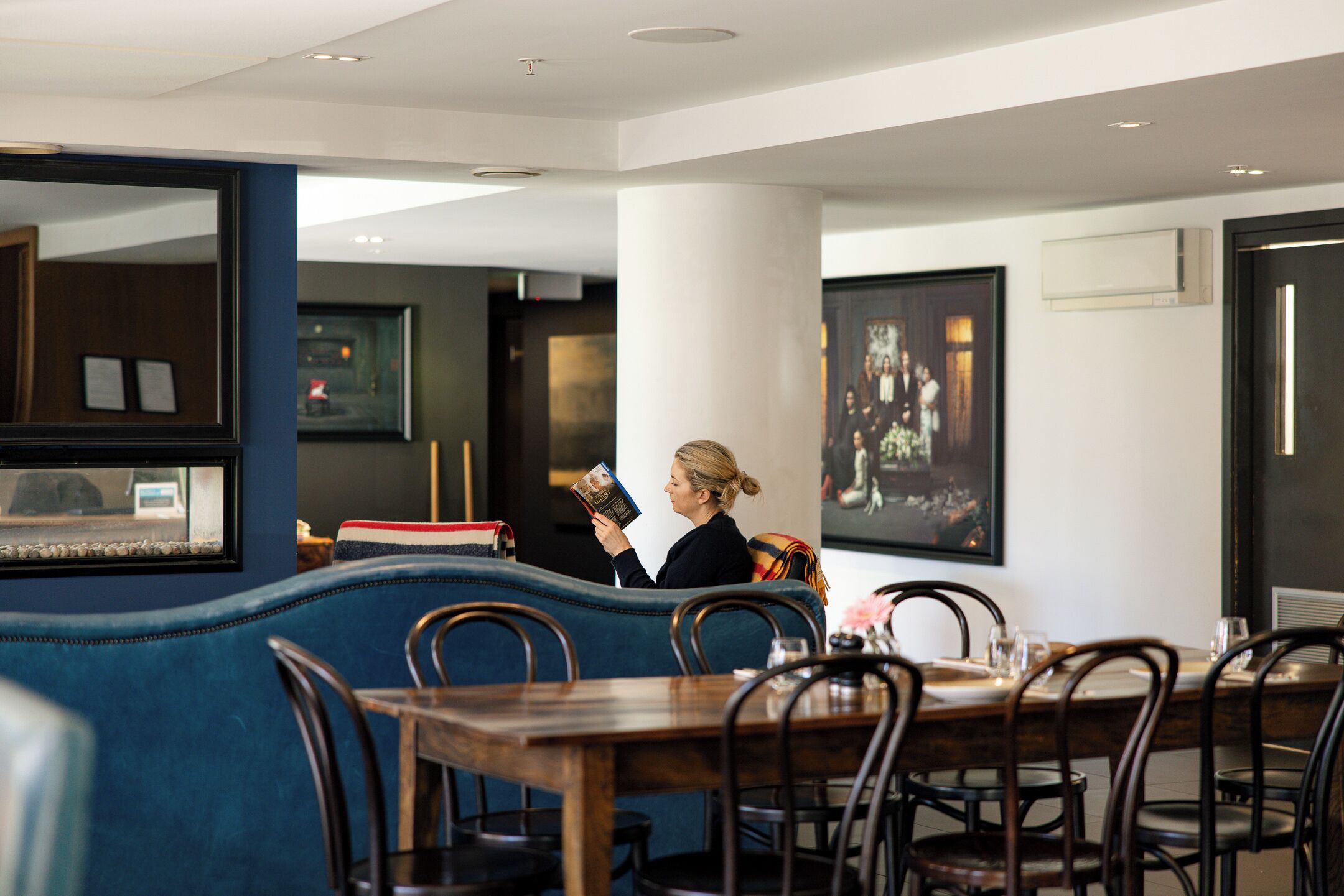 A woman reading a magazine in a modern restaurant with blue seating, wooden tables, and framed artwork on the walls.