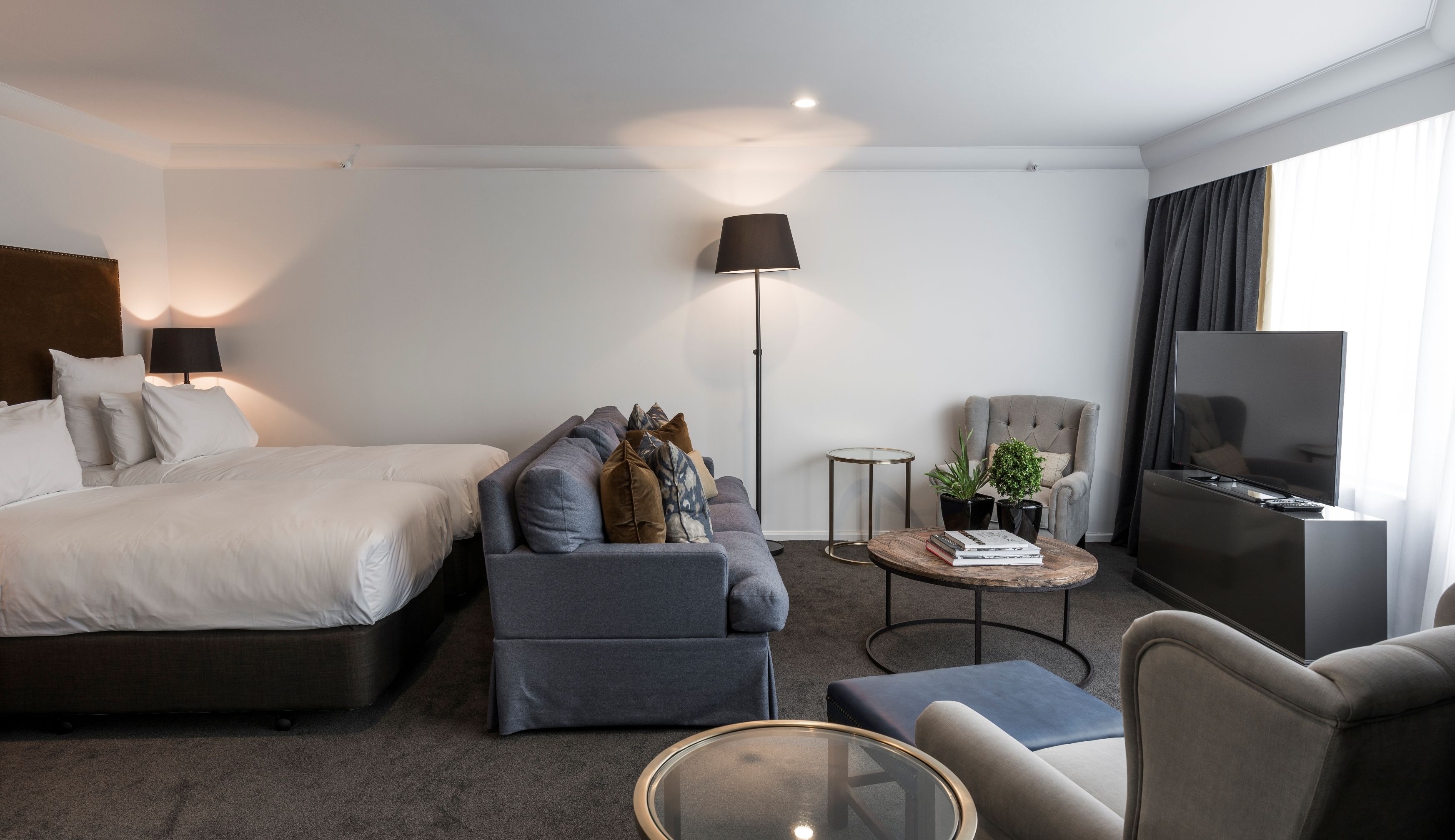 A modern hotel room with a bed, sofa, armchair, coffee table, TV, and a floor lamp.