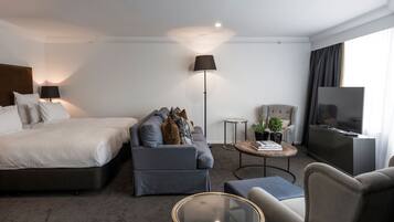 A modern hotel room with a bed, sofa, armchair, coffee table, TV, and a floor lamp.