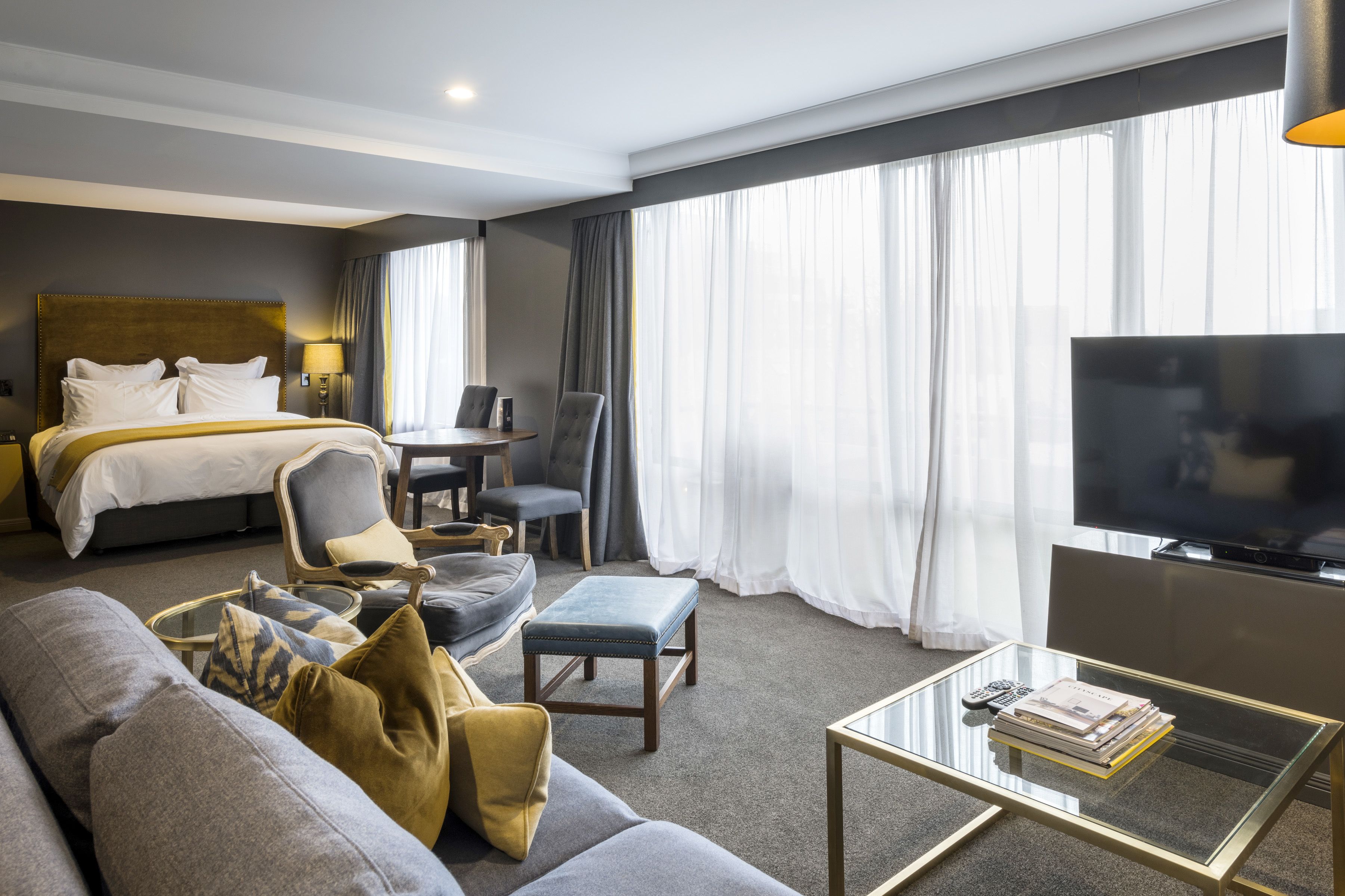 A hotel room with a bed, a sofa, a coffee table, a TV, and a large window with curtains.