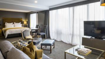 A hotel room with a bed, a sofa, a coffee table, a TV, and a large window with curtains.