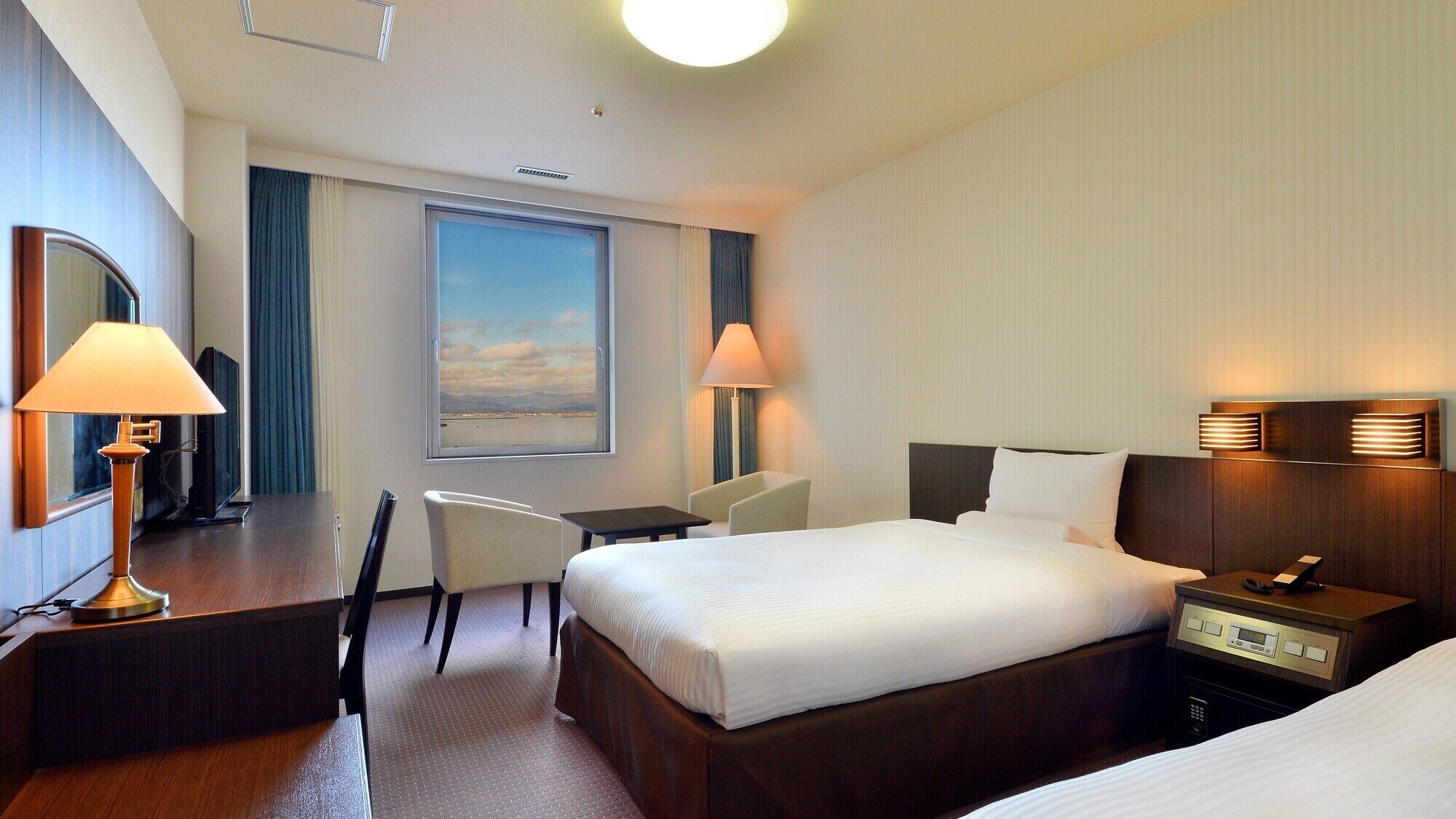 Een hotelkamer met een bed, nachtkastje, lamp, bureau, stoel en een groot raam met uitzicht.