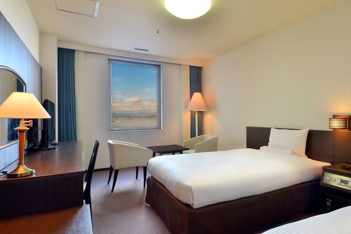 Een hotelkamer met een bed, nachtkastje, lamp, bureau, stoel en een groot raam met uitzicht.
