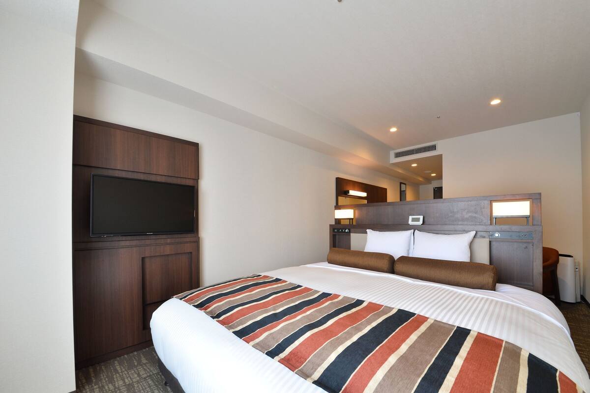 Een hotelkamer met een groot bed, een aan de muur bevestigde televisie en een klein bureau met een lamp.