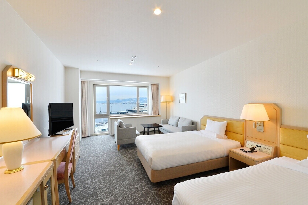 A hotel room with two beds, a desk with a TV, a chair, a small table, and a view of the sea.