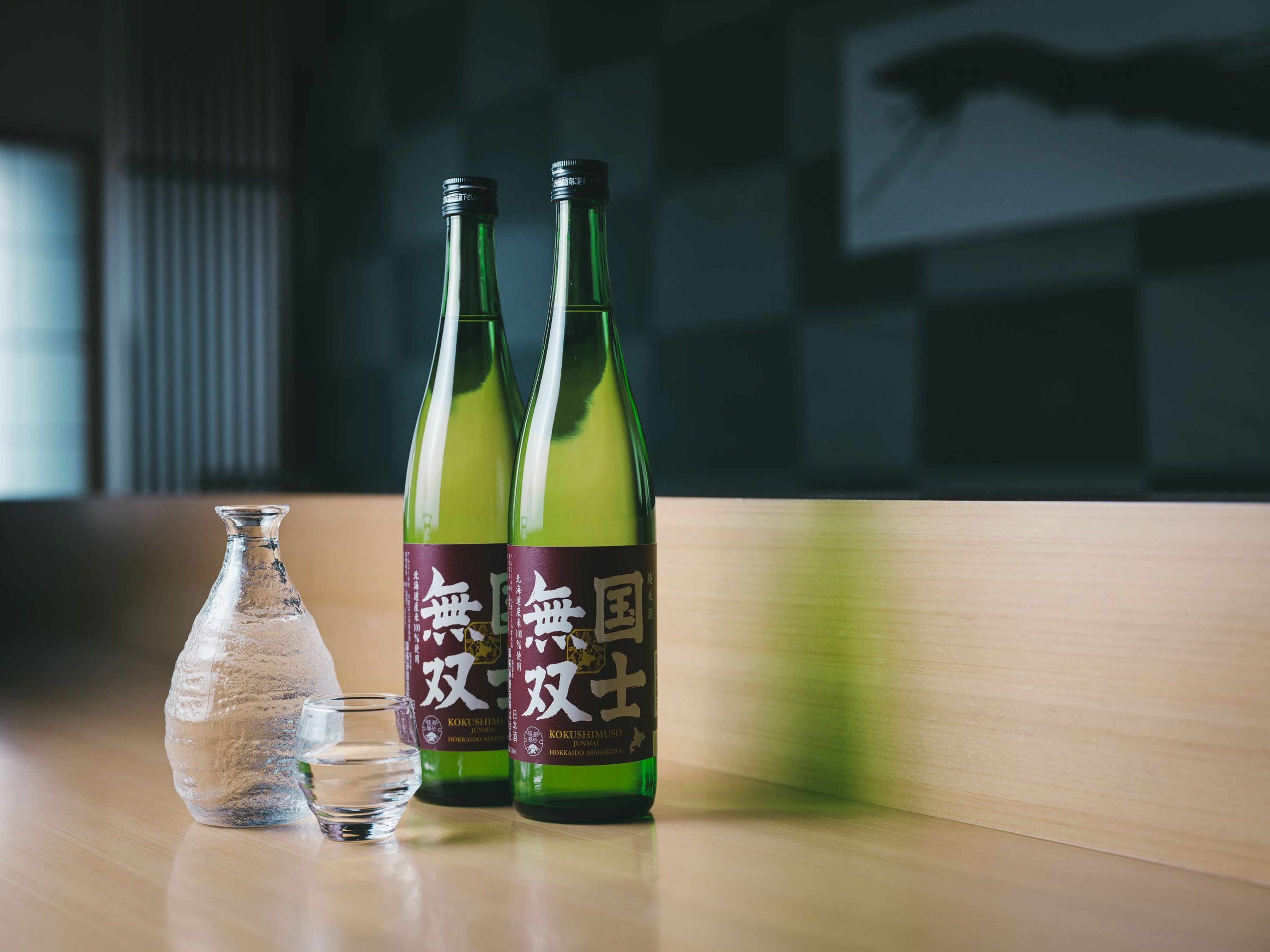 Twee flessen sake met etiketten, een glazen fles met vloeistof, en een klein glas op een houten oppervlak.