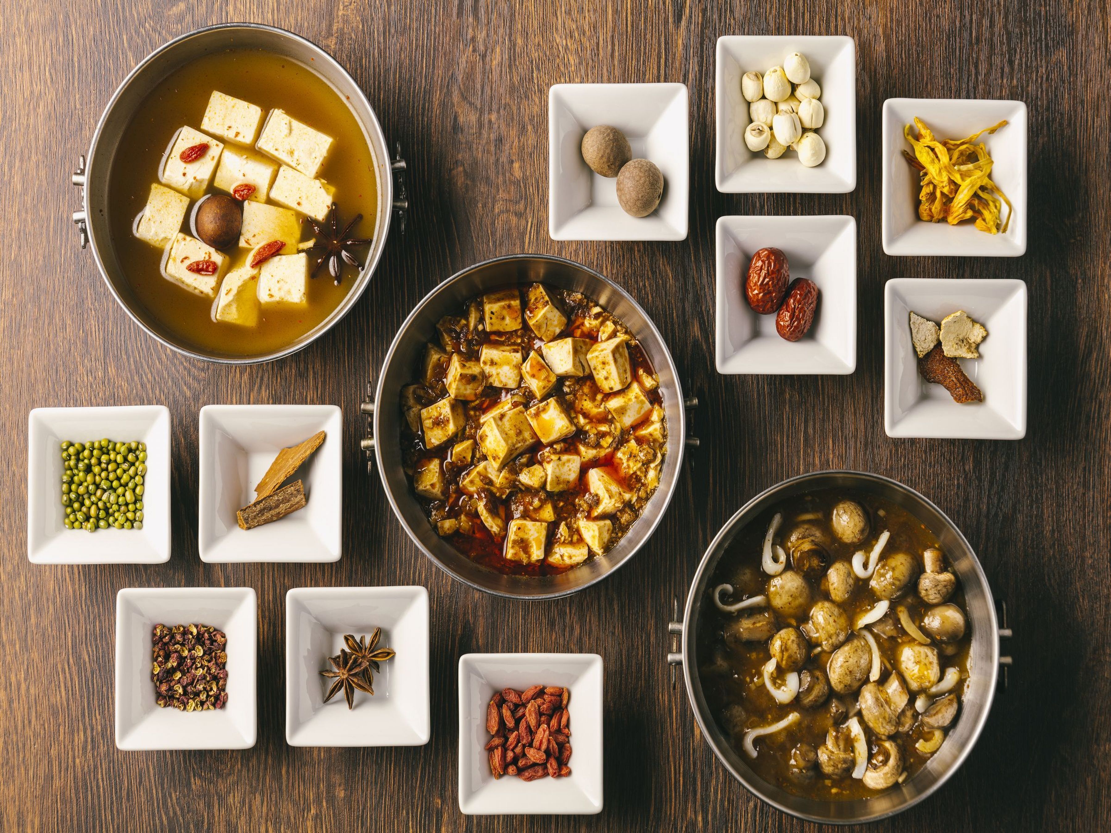 A variety of dishes and ingredients arranged on a wooden table, including tofu, mushrooms, and various herbs and spices.