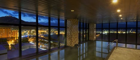 Amplio espacio interior con ventanas grandes que ofrecen vistas a un paisaje urbano y una montaña al atardecer.